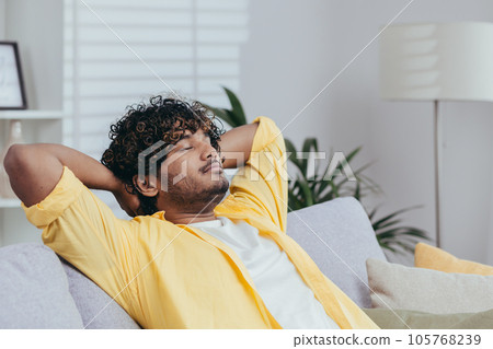 Young man relaxing alone at home on sofa, close-up Hispanic man with hands behind head dozing and dreaming with closed eyes in living room of house. 105768239