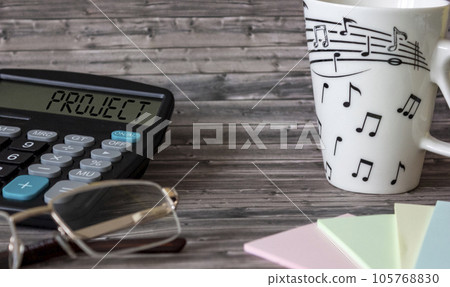 On a wooden table lies a calculator with the text Project, stickers, glasses and a mug 105768830