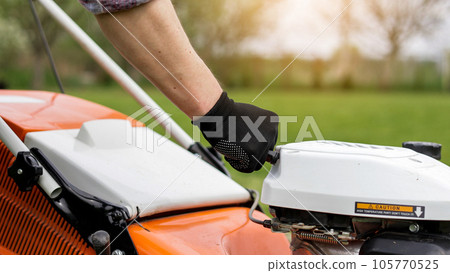 Unrecognisable gardener man in protective gloves starts the lawnmower before cutting green grass lawn in his backyard. Man with motorised lawnmower cares for landscaping lawn 105770525