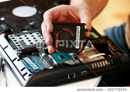 Fixing laptop. Repairman holds cooling fan and repairs from overheating. Disassembling computer, replacement, clean dust and dirt cooling system on notebook. Professional electronic device service Fixing laptop. Repairman holds cooling fan and repairs from overheating. Disassembling computer, replacement, clean dust and dirt cooling system on notebook. Professional electronic device service 105770541