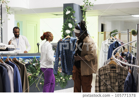 Clothing store customer in headphones holding shirt on hanger to try on in fitting room. Stylish african american man choosing outfit while listening to music playlist in earphones in boutique Clothing store customer in headphones holding shirt on hanger to try on in fitting room. Stylish african american man choosing outfit while listening to music playlist in earphones in boutique 105771378