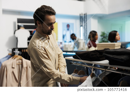 Customer browsing rack with apparel while selecting trendy casual outfit in clothing store. Man exploring hanger with garment while shopping in retail department boutique Customer browsing rack with apparel while selecting trendy casual outfit in clothing store. Man exploring hanger with garment while shopping in retail department boutique 105771416