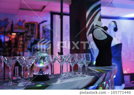 Glasses for alcohol drink and disco ball on bar counter for discotheque party in nightclub with no people. Booze beverages prepared for music concert in dark club with spotlights glow 105771440