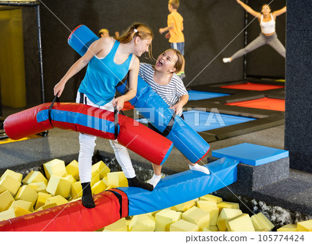 Smiling positive girls in sportswear having funny wrestling by inflatable logs in indoor game trampoline club 105774424
