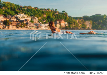 Surfer woman rowing on surfboard in ocean with coastline landscape on background. Surfer woman rowing on surfboard in ocean with coastline landscape on background. 105780265