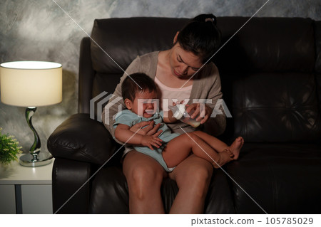 stressed mother trying to feeding milk bottle to crying infant baby on sofa in the living room at night stressed mother trying to feeding milk bottle to crying infant baby on sofa in the living room at night 105785029