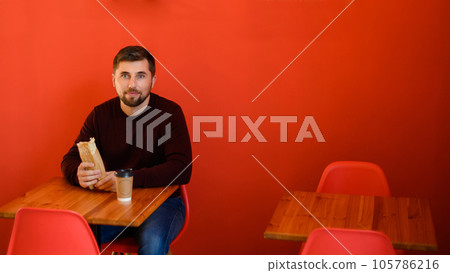 A man with a beard sits in a fast food restaurant and holds a shawarma in his hand and has a glass of coffee. 105786216