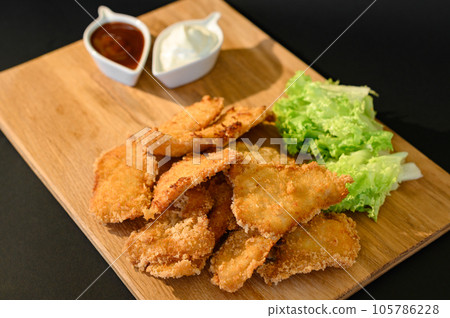 Chicken nuggets with sauces, mayonnaise and ketchup, on a wooden board on a black background and salad leaves. Chicken nuggets with sauces, mayonnaise and ketchup, on a wooden board on a black background and salad leaves. 105786228