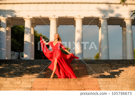 A woman in a long red dress against the backdrop of sunrise, bright golden light of the sun's rays. The concept of femininity, harmony. 105786673
