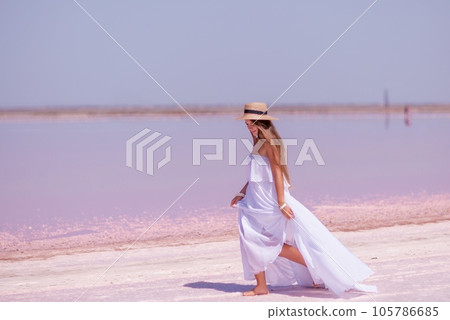 Woman in pink salt lake. She in a white dress and hat enjoys the scenic view of a pink salt lake as she walks along the white, salty shore, creating a lasting memory. 105786685