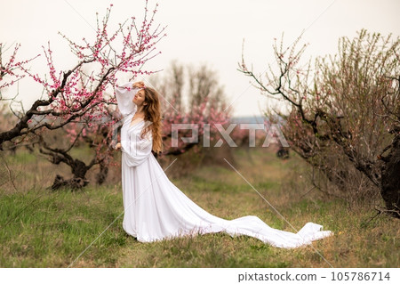 Woman peach blossom. Happy curly woman in white dress walking in the garden of blossoming peach trees in spring 105786714