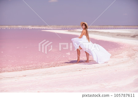 Woman in pink salt lake. She in a white dress and hat enjoys the scenic view of a pink salt lake as she walks along the white, salty shore, creating a lasting memory. 105787611