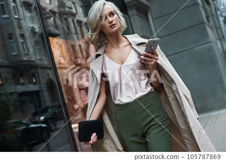 Fashion. Young stylish woman walking on the city street browsing internet on smartphone looking aside pensive 105787869