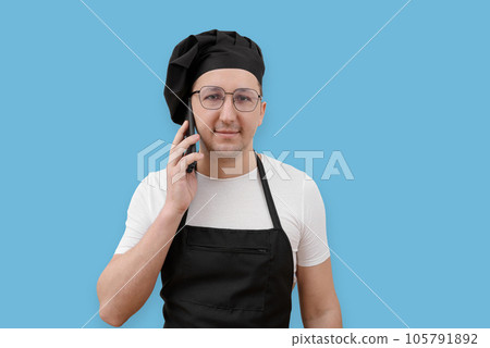 Young chef talking on the phone and looking at the camera on a blue background. The concept of cooking Young chef talking on the phone and looking at the camera on a blue background. The concept of cooking 105791892