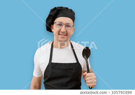 Portrait of a cook in an apron and with a spoon in his hand looking at the camera on a blue background. Portrait of a cook in an apron and with a spoon in his hand looking at the camera on a blue background. 105791899