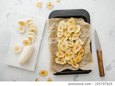 Tray with dried banana chips and raw banana with kitchen knife.Top view. 105797829