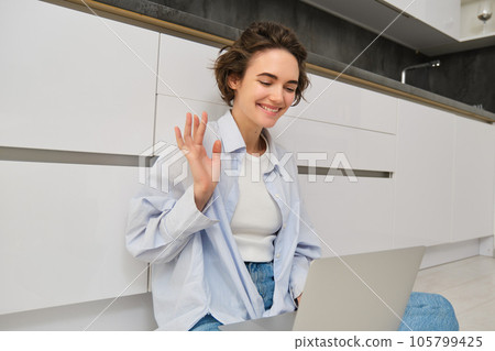 Friendly girl talks on laptop, connects to work online meeting via computer, sits on floor and waves hand at camera Friendly girl talks on laptop, connects to work online meeting via computer, sits on floor and waves hand at camera 105799425