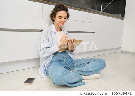 Thoughtful young woman, creative artist draws in her notebook, makes sketches, writes down in her journal diary, sits on floor and thinks Thoughtful young woman, creative artist draws in her notebook, makes sketches, writes down in her journal diary, sits on floor and thinks 105799471