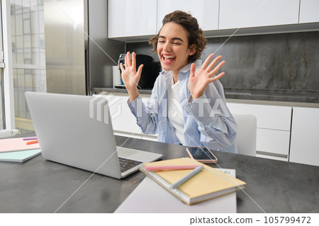 Beautiful girl waves hands at laptop camera, says hello to coworkers, connects to work meeting online, studies on remote from her kitchen 105799472