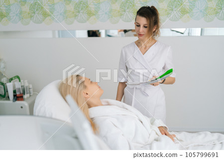 Portrait of female doctor take notes while discussing with young woman client lying on couch before rf-lifting procedure in modern light beauty clinic. Concept of non-surgery cosmetology. 105799691