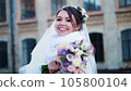 portrait of a beautiful bride, in a white dress, veil and fur coat. bride is smiling happily, holding a wedding bouquet. against the background of ancient architecture, winter wedding. 105800104