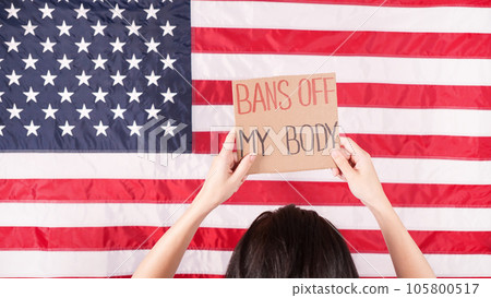 Young woman protester holds cardboard with Bans Of My Body signs against USA flag on background. Girl protesting against anti-abortion laws. Womens rights freedom. Young woman protester holds cardboard with Bans Of My Body signs against USA flag on background. Girl protesting against anti-abortion laws. Womens rights freedom. 105800517