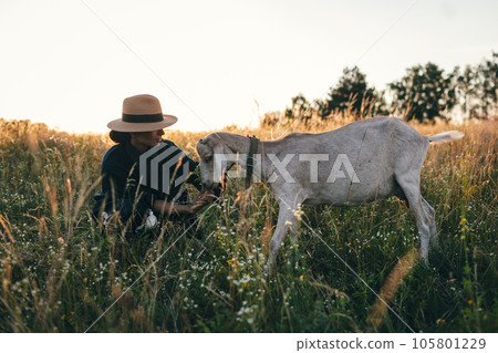 Young woman in the field is grazing her white goats. The girl and goats in the meadow in summer. Love for animals. Goat farm. Pets. Happy woman with animal. Kindness and love for animals. Kisses a pet 105801229