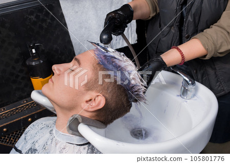 The hands of a girl, an experienced salon worker in black gloves, wash off the paint from the hair of a young guy's client with water and rain over the sink in the process of dyeing hair 105801776