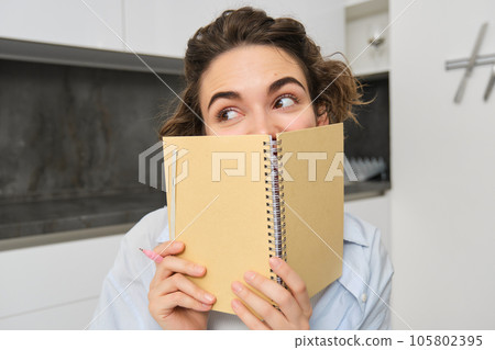 Portrait of cute brunette girl, looks away, hides her face behind planner, notebook, gets creative and writes in her journal 105802395