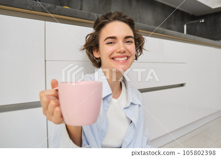 Positive, smiling young woman sits on kitchen floor, extends hand with cup, offers you mug with coffee or tea, enjoys her drink Positive, smiling young woman sits on kitchen floor, extends hand with cup, offers you mug with coffee or tea, enjoys her drink 105802398