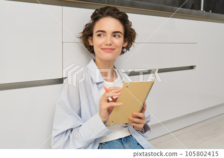 Portrait of cute brunette woman sits on floor, draws in her planner, writes diary on floor in kitchen and smiles Portrait of cute brunette woman sits on floor, draws in her planner, writes diary on floor in kitchen and smiles 105802415