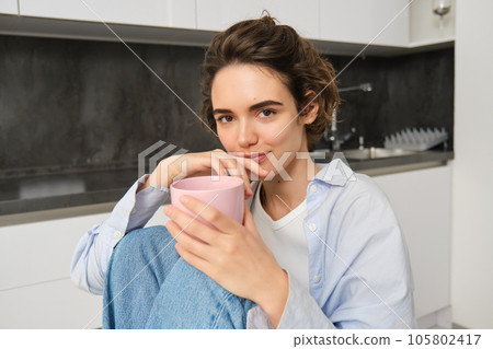 Close up portrait of woman sitting at home with cup of aromatic coffee, enjoying hot drink on weekend, smiling at camera Close up portrait of woman sitting at home with cup of aromatic coffee, enjoying hot drink on weekend, smiling at camera 105802417