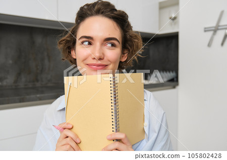 Portrait of smiling brunette girl, holds her planner, writes in notebook and looks happy, laughs, does homework, makes notes, sits at home in kitchen 105802428