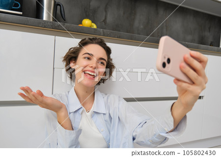 Portrait of young woman video chats, records vlog while sits on kitchen floor. Cute girl takes selfie on smartphone 105802430