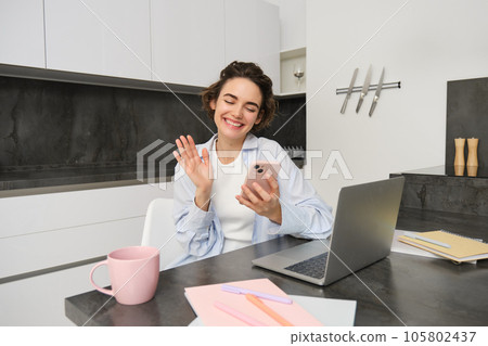 Portrait of young woman video chats with friend on smartphone, waves hand at mobile phone camera, calls someone using application 105802437