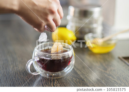 Womans hand puts the tea bag in the glass cup with hot water 105815575