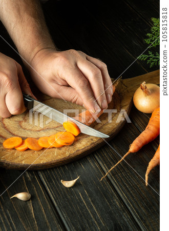 The cook cuts raw carrots with a knife on a wooden cutting board for cooking. Peasant food. The cook cuts raw carrots with a knife on a wooden cutting board for cooking. Peasant food. 105815818