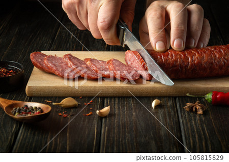 The cook cuts homemade sausage on a wooden cutting board. Cooking delicious sandwiches for a snack at home The cook cuts homemade sausage on a wooden cutting board. Cooking delicious sandwiches for a snack at home 105815829