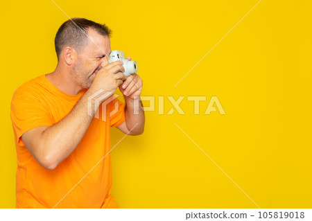 Happy bearded hispanic man in a t-shirt smiling and taking profile picture with a small photo camera against a yellow background. 105819018