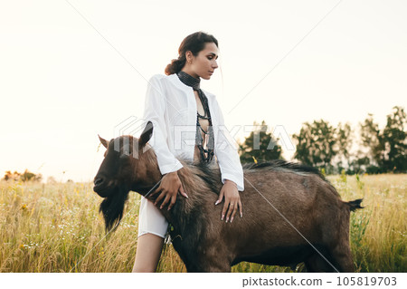 Young woman in the field is walking with goats. The girl and goats in the meadow in summer. Love for animals. Goat farm. Pets. Happy woman with animal. Kindness and love for animals. Kisses a pet Young woman in the field is walking with goats. The girl and goats in the meadow in summer. Love for animals. Goat farm. Pets. Happy woman with animal. Kindness and love for animals. Kisses a pet 105819703