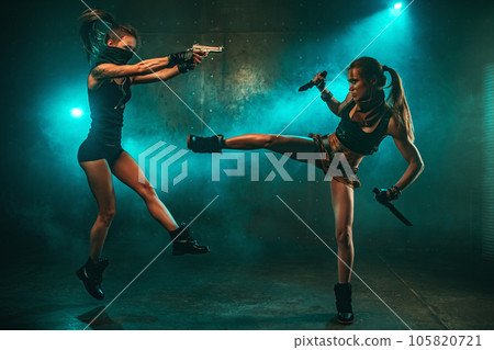 Two women with guns and knifes fighting in industrial interior with smoke, tattoo on body 105820721