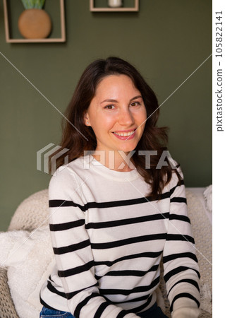 Young attractive caucasian woman sits alone on the sofa in the living room during the day and smiles cutely while looking at the camera Young attractive caucasian woman sits alone on the sofa in the living room during the day and smiles cutely while looking at the camera 105822141