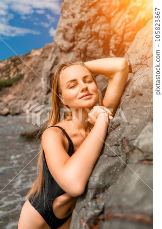 Woman travel sea. Attractive blonde woman in a black swimsuit enjoying the sea air on the seashore around the rocks. Travel and vacation concept. 105823867