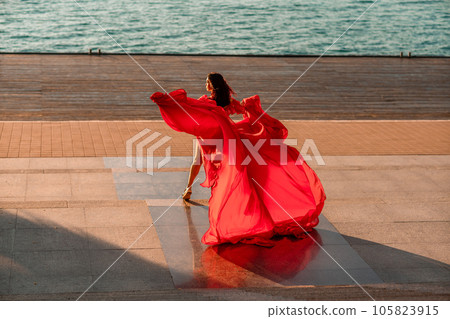 Sunrise red dress. A woman in a long red dress against the backdrop of sunrise, bright golden light of the sun's rays. The concept of femininity, harmony. 105823915