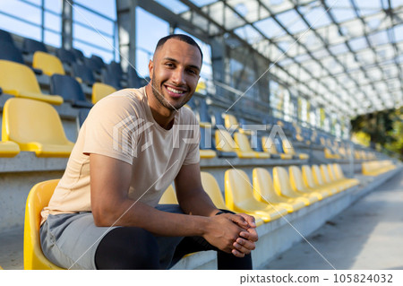 Portrait of young athlete in stadium, man sitting on chair smiling and looking at camera close up, runner resting from training and fitness. Portrait of young athlete in stadium, man sitting on chair smiling and looking at camera close up, runner resting from training and fitness. 105824032