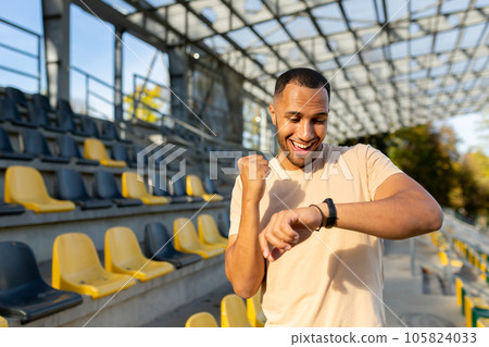 Happy and successful man satisfied with the results of physical training celebrating good results of fitness and running looking at smart watch fitness bracelet walking in the air in the stadium. 105824033