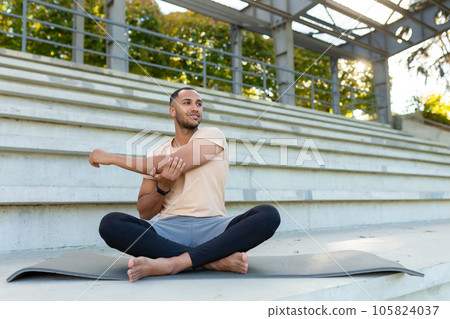 A young African American man is playing sports at the stadium. Sits on a mat, performs an exercise, stretches, does yoga. 105824037