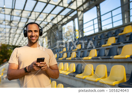 Sportsman using telephone smiling and looking at camera, man on morning jog and exercising at sports stadium, man listening to music in headphones and podcasts online radio. Sportsman using telephone smiling and looking at camera, man on morning jog and exercising at sports stadium, man listening to music in headphones and podcasts online radio. 105824043