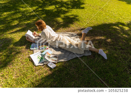 An adult relaxed woman lying on the blanket in the park and drawing in her sketchbook An adult relaxed woman lying on the blanket in the park and drawing in her sketchbook 105825238