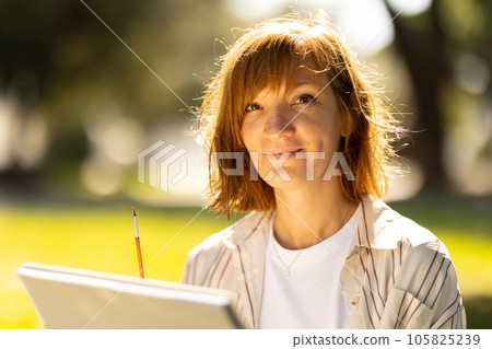 Adult woman artist sitting in the park holding a sketchbook Adult woman artist sitting in the park holding a sketchbook 105825239